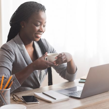 Coffee break. Successful black businesswoman holding cup with hot drink at workplace and looking at laptop screen, copy space